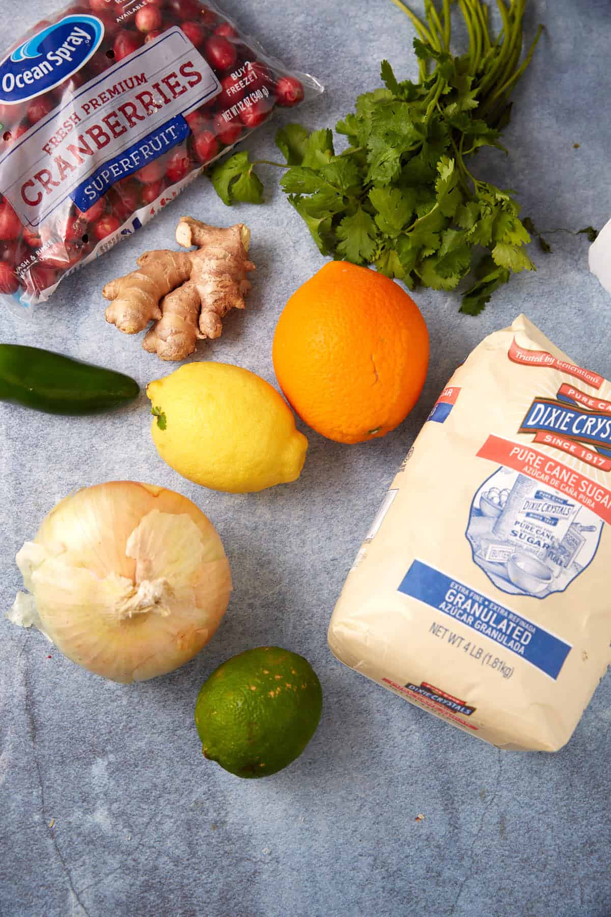 A collection of fresh ingredients on a blue surface, including a bag of cranberries, cilantro, ginger, jalapeño, lemon, orange, lime, a yellow onion, and a bag of granulated sugar.