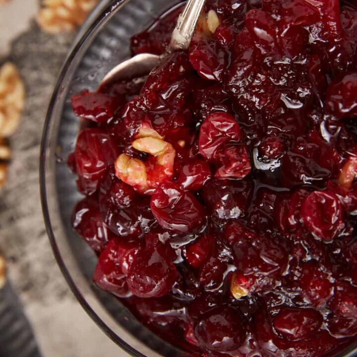 A bowl of chunky homemade cranberry sauce with visible walnut pieces, served with a silver spoon. Walnuts are scattered on a cloth napkin in the background.