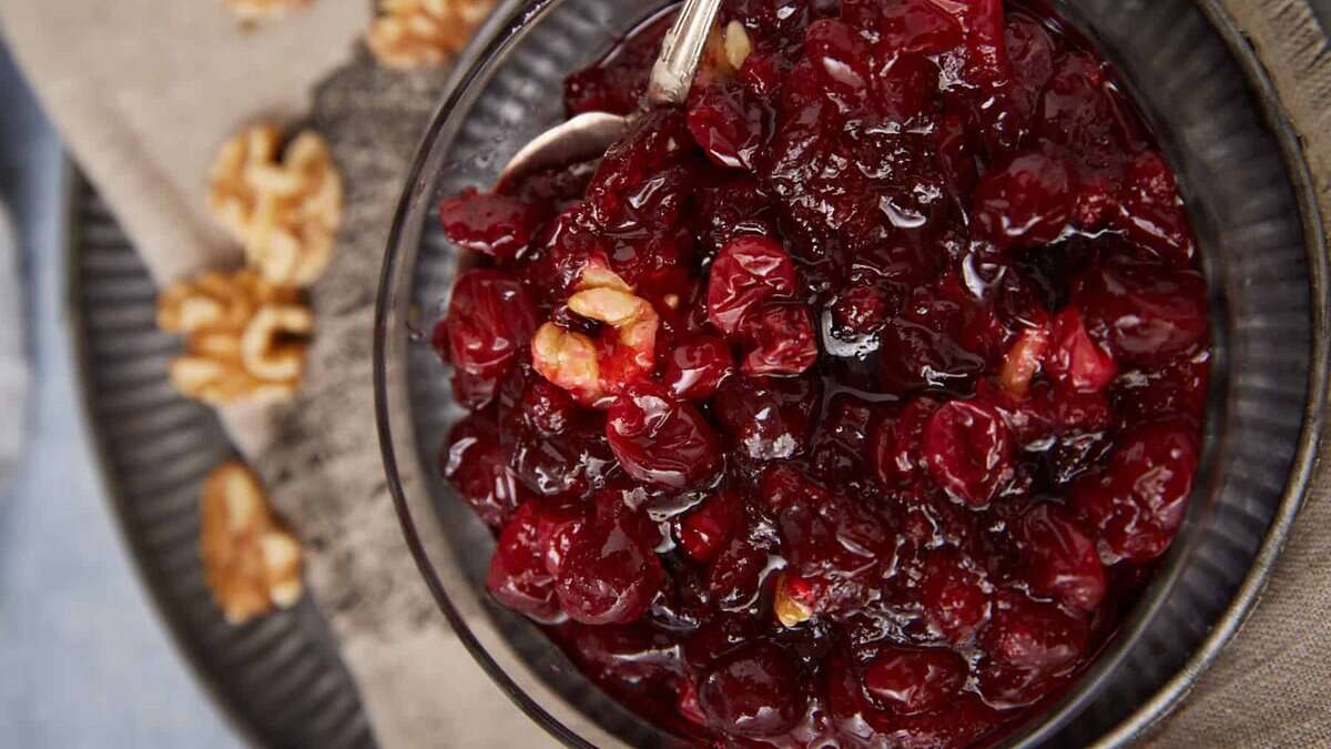 A bowl of chunky homemade cranberry sauce with visible walnut pieces, served with a silver spoon. Walnuts are scattered on a cloth napkin in the background.