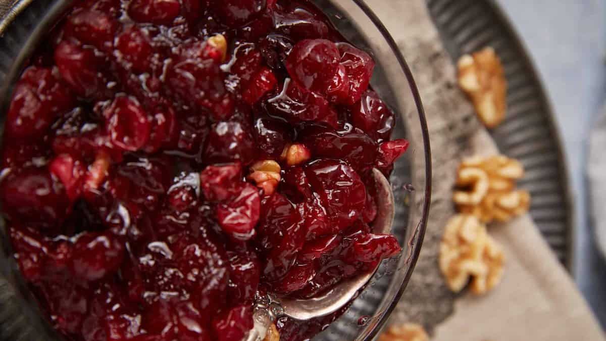 A close-up of a bowl filled with homemade cranberry sauce, mixed with visible pieces of walnut, and a spoon resting inside. In the background, whole walnut pieces are scattered on a plate.