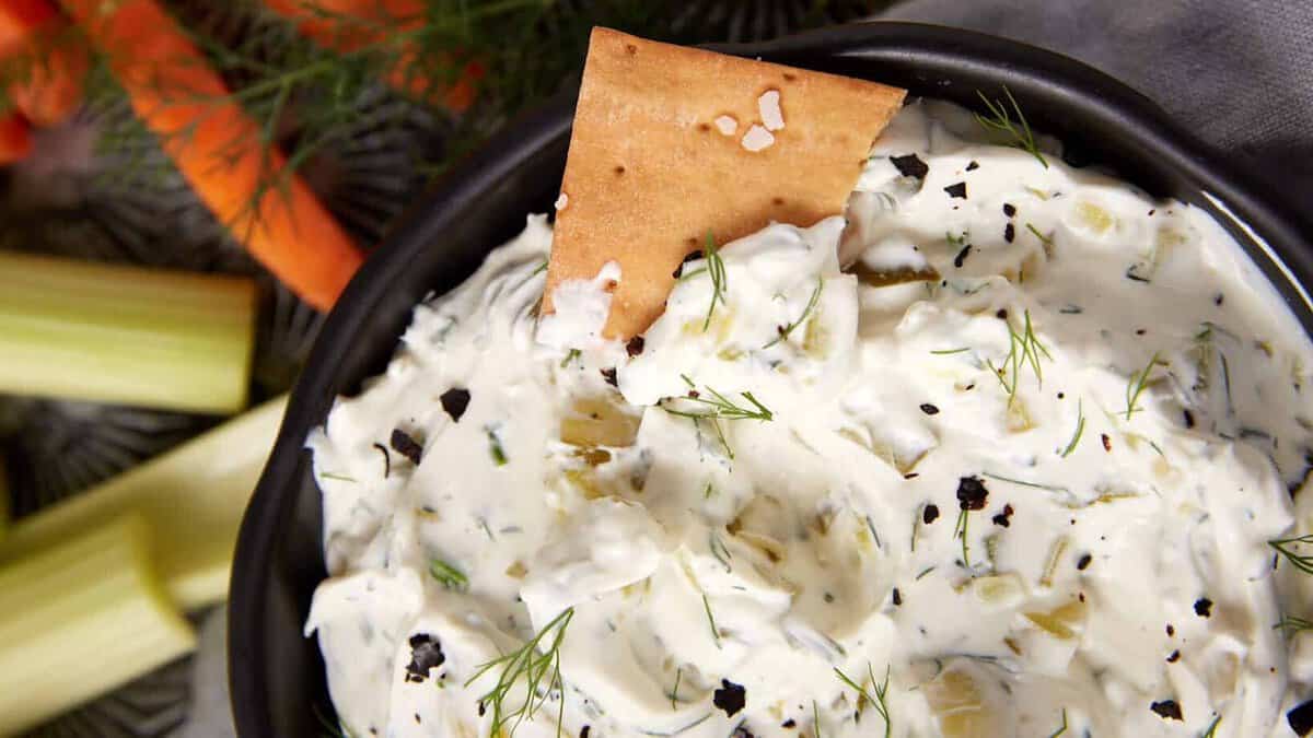 A bowl of creamy dill pickle dip garnished with dill and cracked pepper, with a cracker dipped in it. Surrounding the bowl are celery sticks, carrot sticks, fresh dill, and crackers.