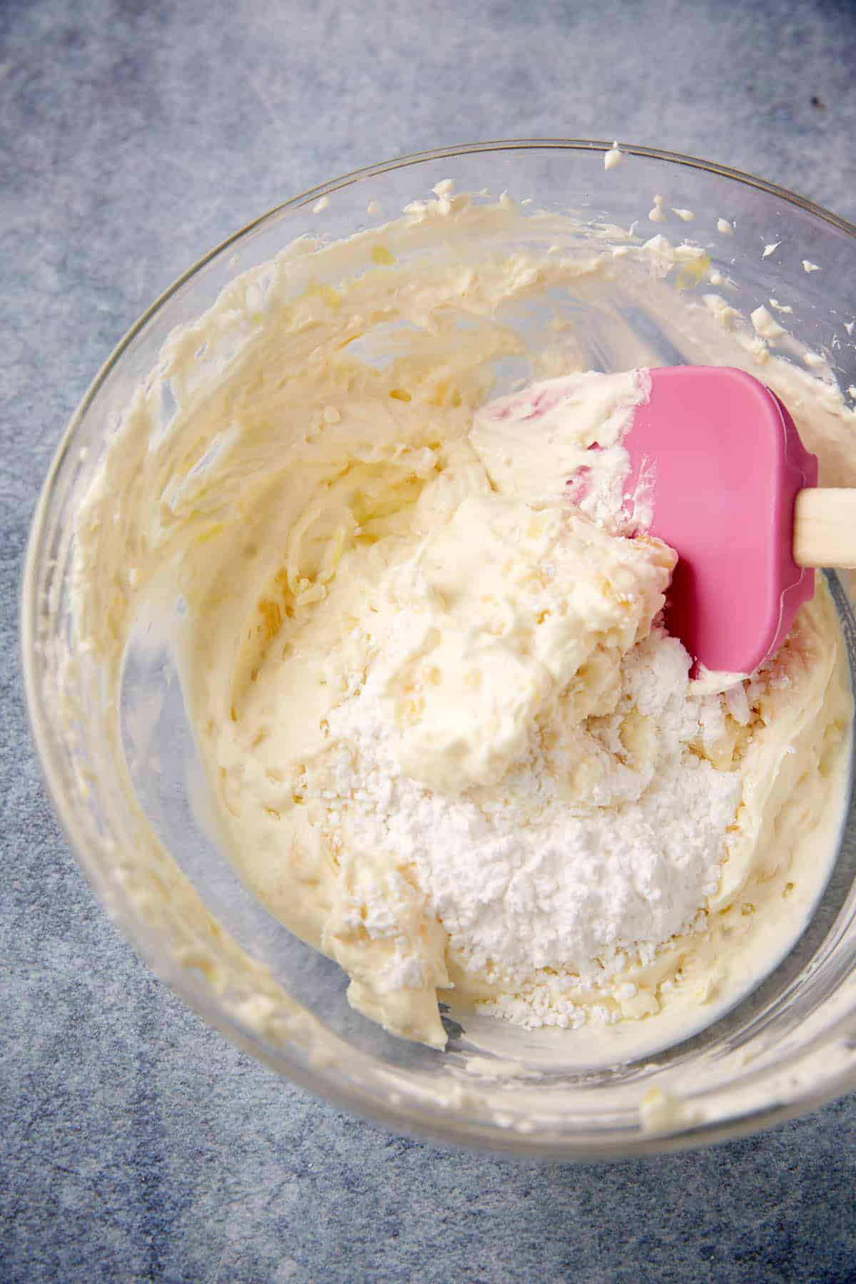 A glass bowl containing a creamy mixture and a mound of powdered sugar, being mixed with a pink silicone spatula on a gray countertop.