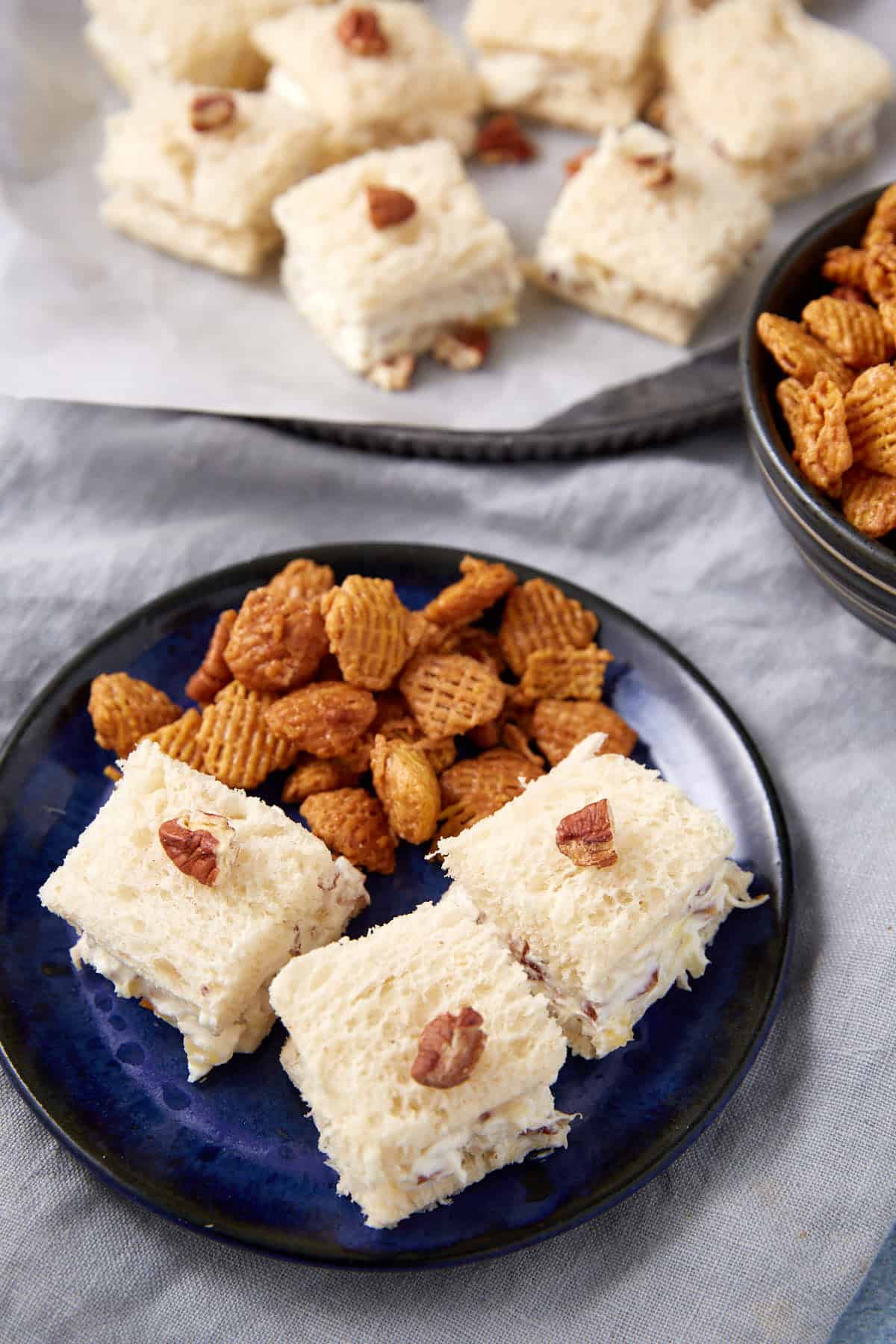 Three square tea sandwiches topped with pecans and some crunchy snack pieces are served on a blue plate. More sandwiches and snacks are seen in the background.