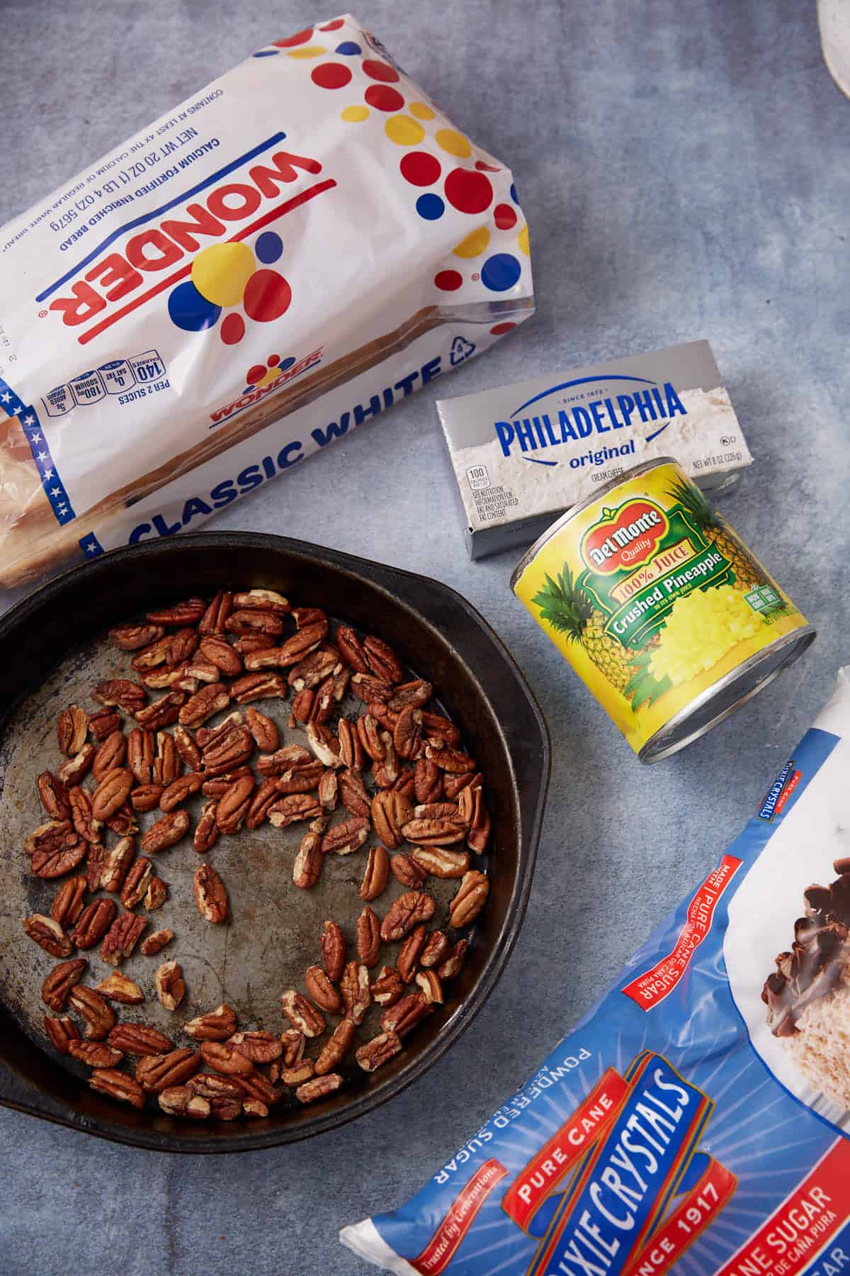 A loaf of Wonder classic white bread, a block of Philadelphia cream cheese, a can of crushed pineapple, a bag of Dixie Crystals sugar, and a skillet with pecans on a blue surface.