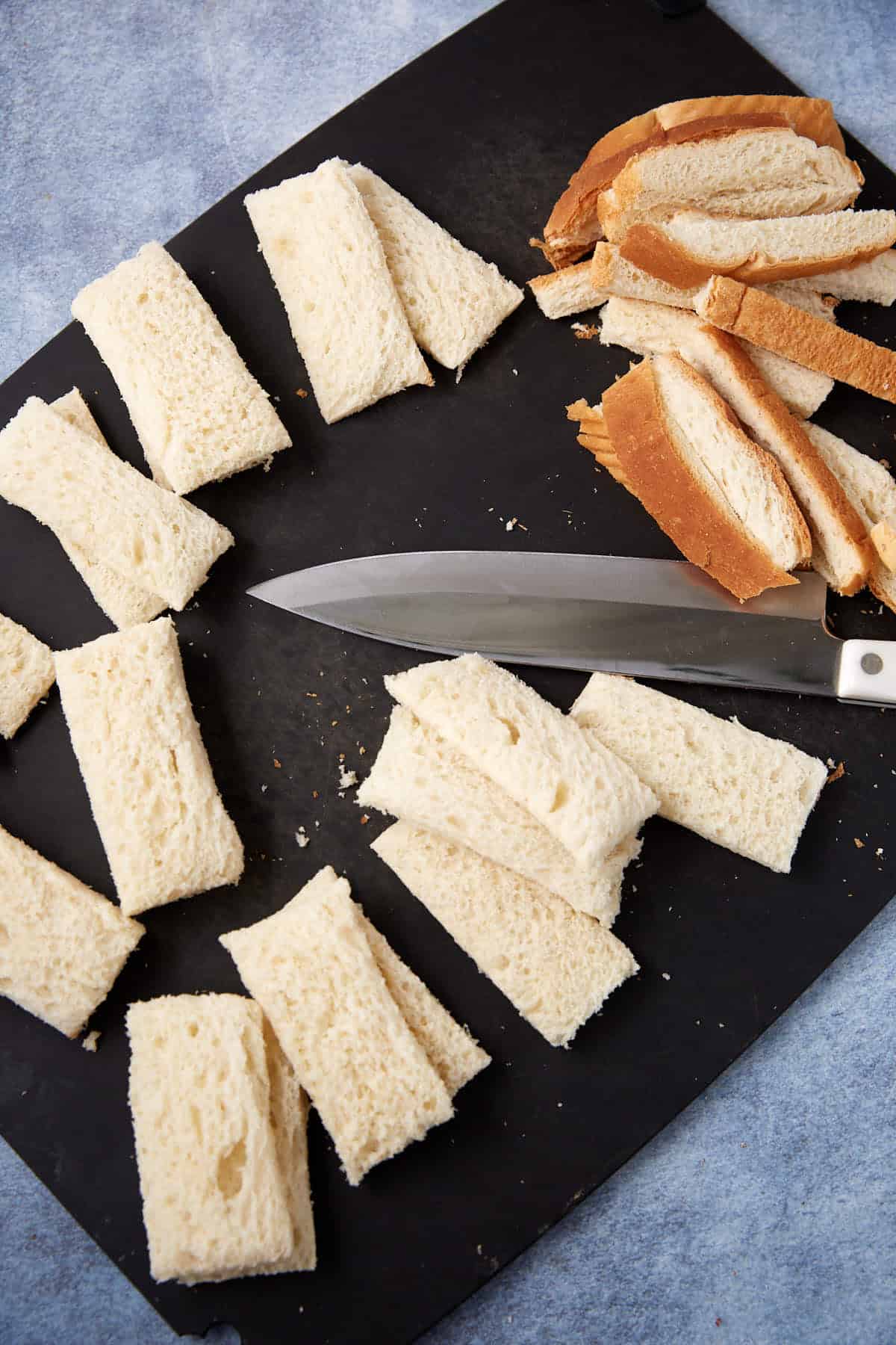 A knife on a black cutting board with crustless white bread rectangles on one side and removed brown crust strips on the other side.