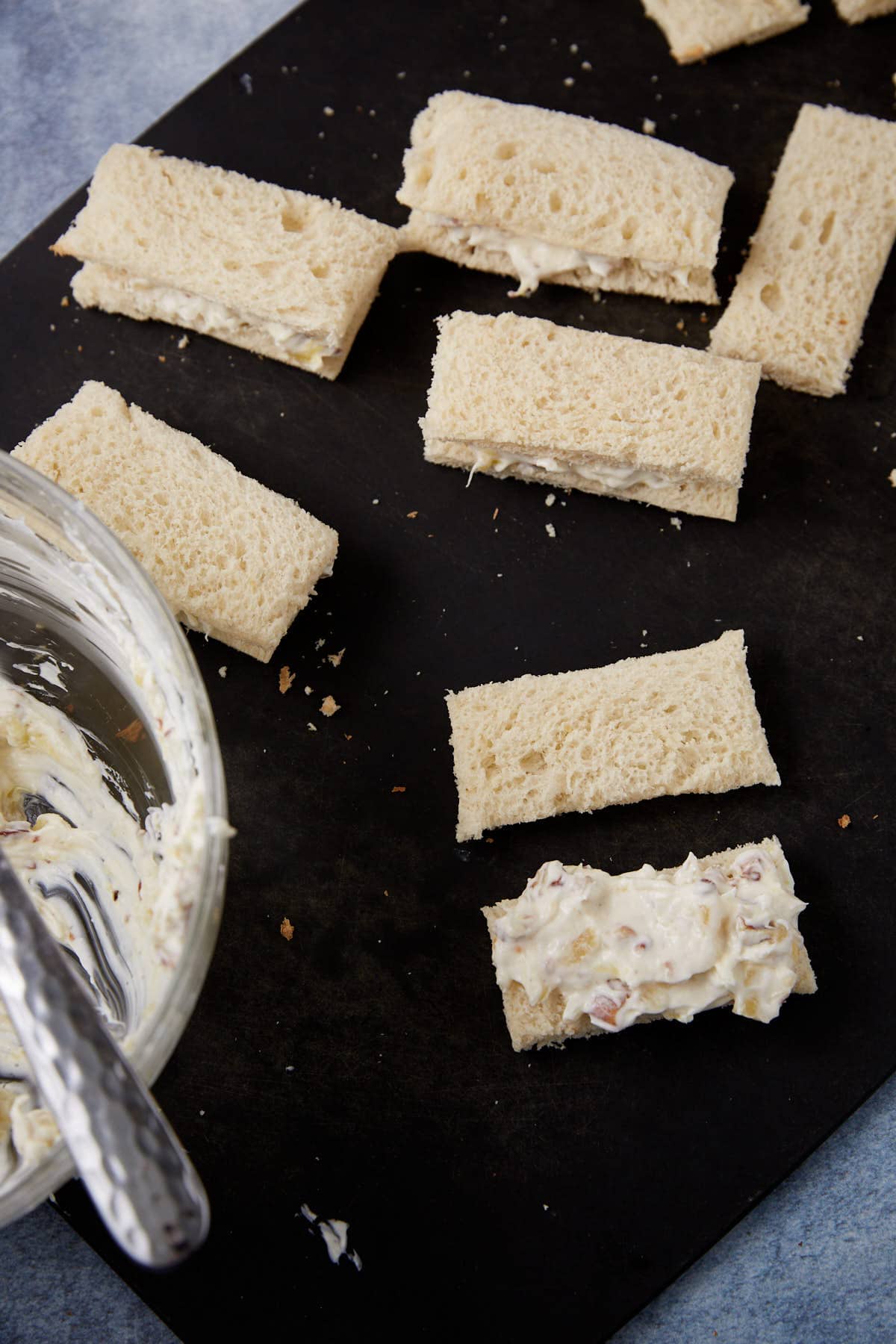Rectangular pieces of white bread are spread with a creamy mixture, next to a bowl containing more of the spread, all on a dark surface.
