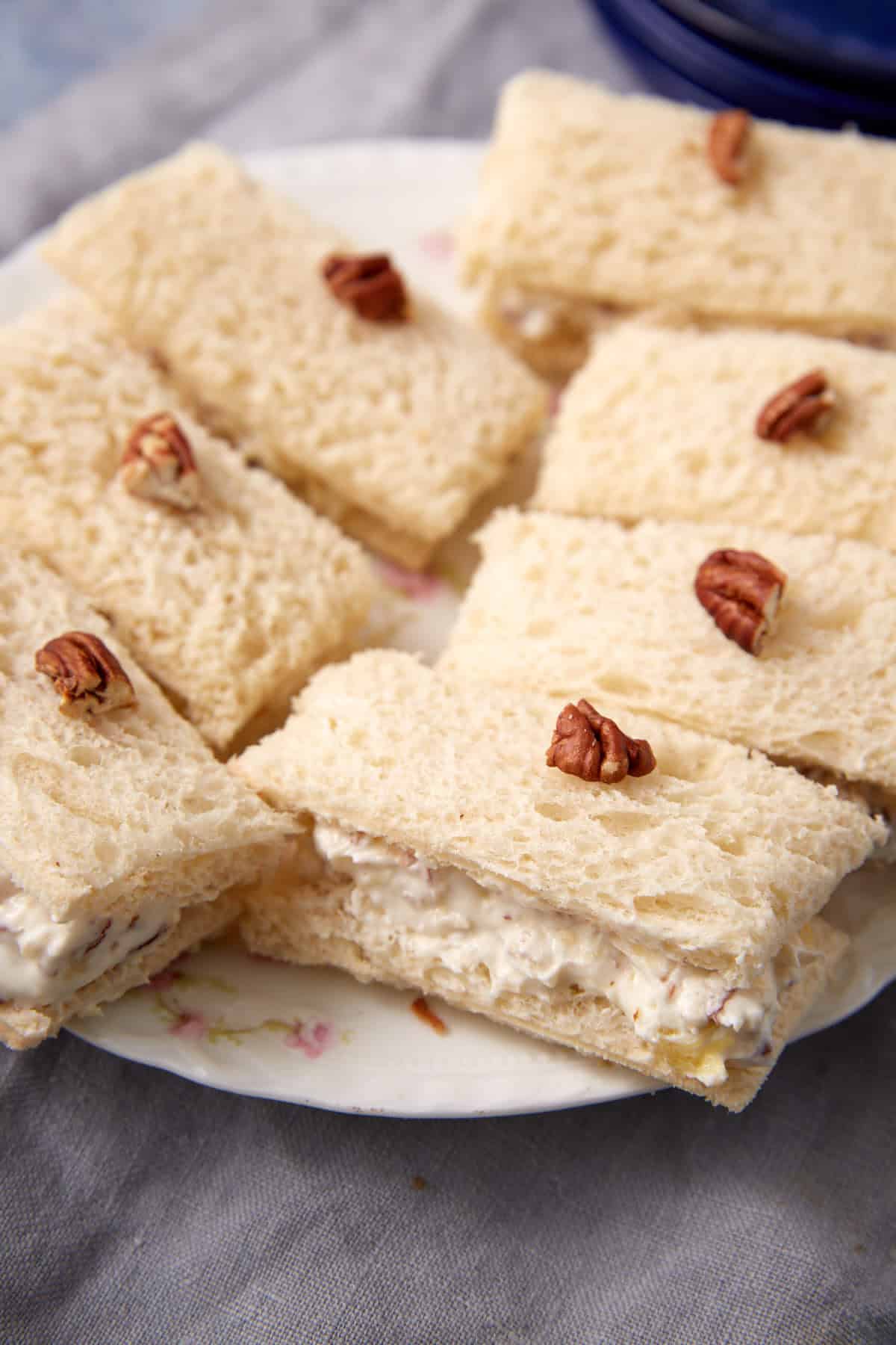 Rectangular tea sandwiches with a creamy filling are arranged on a plate, each topped with a pecan half. The sandwiches are made with soft white bread and served on a floral-patterned dish.