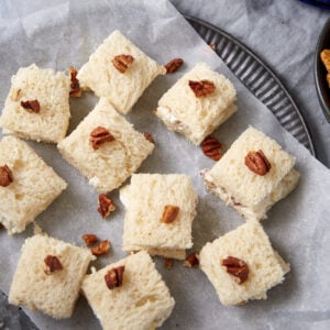 Squares of white bread topped with chopped pecans are arranged on parchment paper on a round tray. Some of the squares are slightly separated, showing a creamy filling inside.