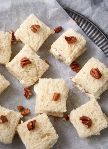 Squares of white bread topped with chopped pecans are arranged on parchment paper on a round tray. Some of the squares are slightly separated, showing a creamy filling inside.