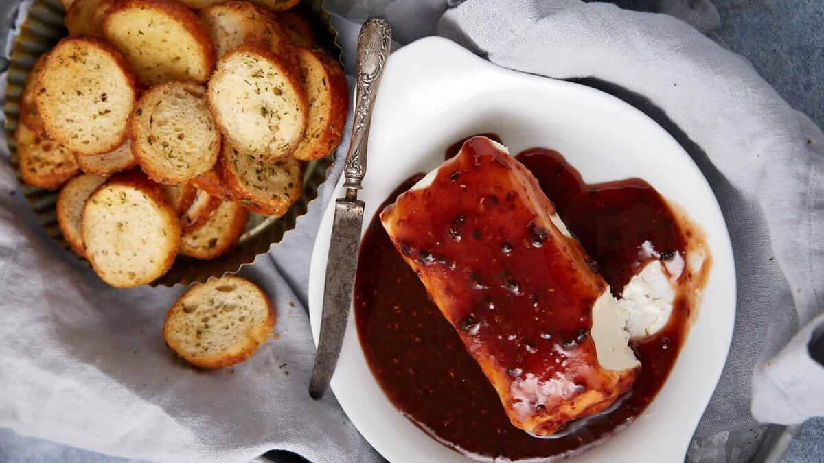 A white dish with a block of goat cheese covered in dark berry sauce sits next to a knife. Behind it, a tray is filled with round toasted bread slices, perfect for enjoying with this raspberry cream cheese dip.
