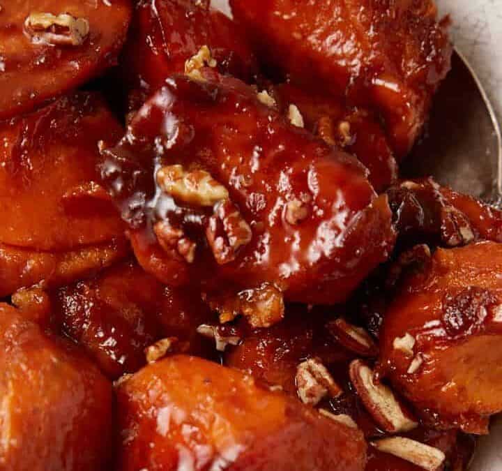 A bowl of glazed sweet potatoes garnished with chopped pecans. The sweet potatoes are caramelized and served in a rustic, white ceramic bowl placed on a gray cloth. A spoon is partially visible in the bowl.