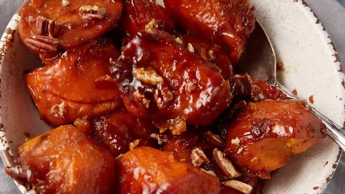 A bowl of glazed sweet potatoes garnished with chopped pecans. The sweet potatoes are caramelized and served in a rustic, white ceramic bowl placed on a gray cloth. A spoon is partially visible in the bowl.
