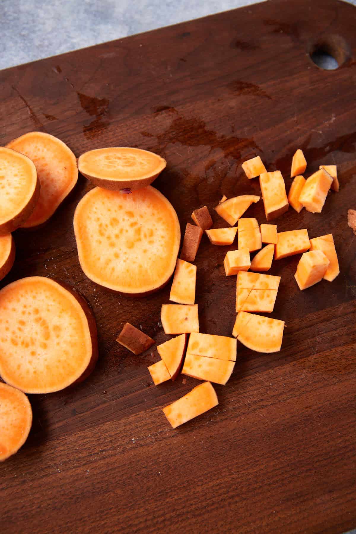 Sliced and diced sweet potatoes on a dark wooden cutting board, with both round slices and small cubes perfect for Sweet Potato Hash.
