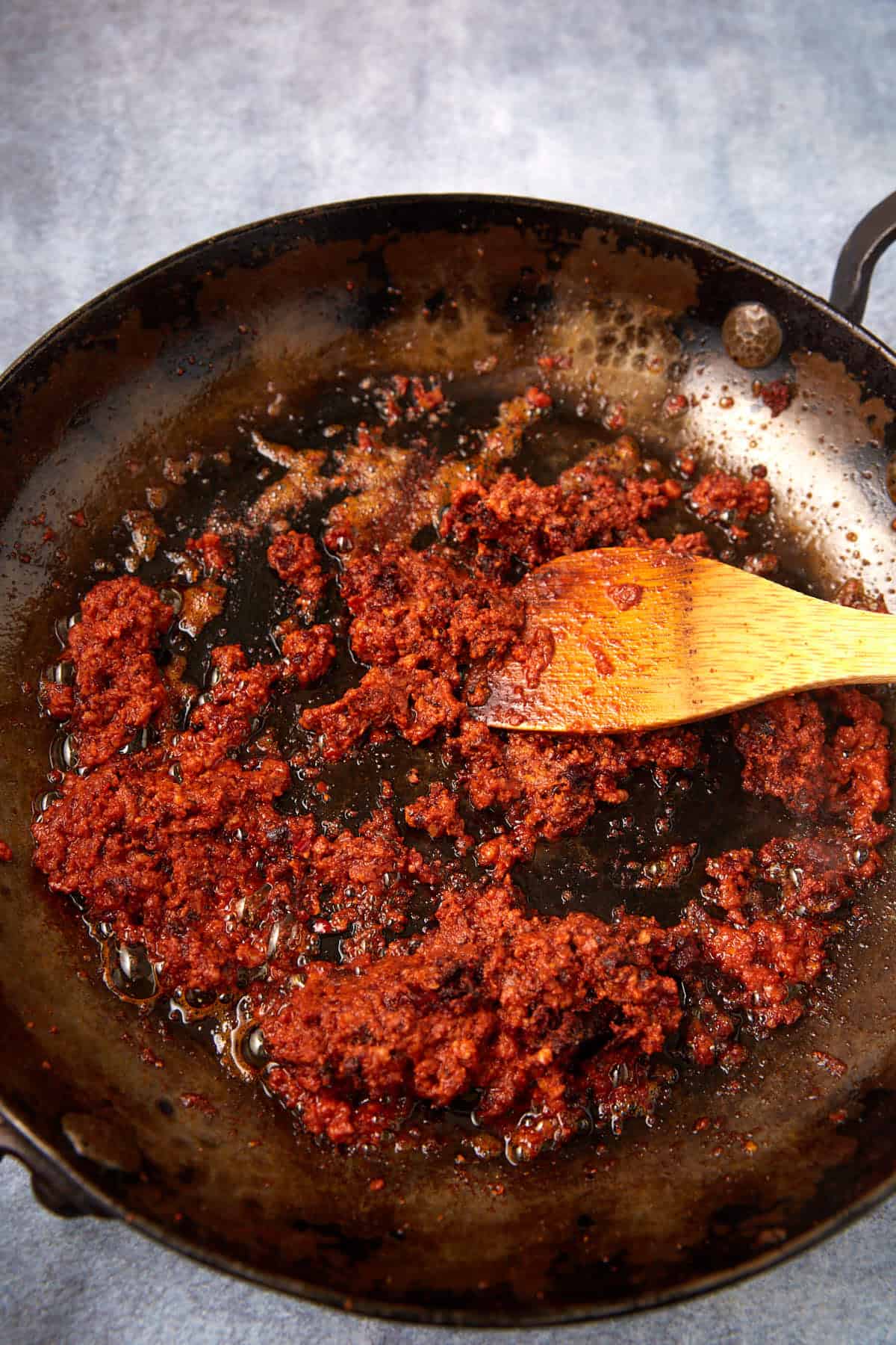 A close-up of a black skillet containing red-brown cooked paste, being stirred with a wooden spatula. The mixture, resembling sweet potato hash, appears oily and is spread across the surface of the pan.