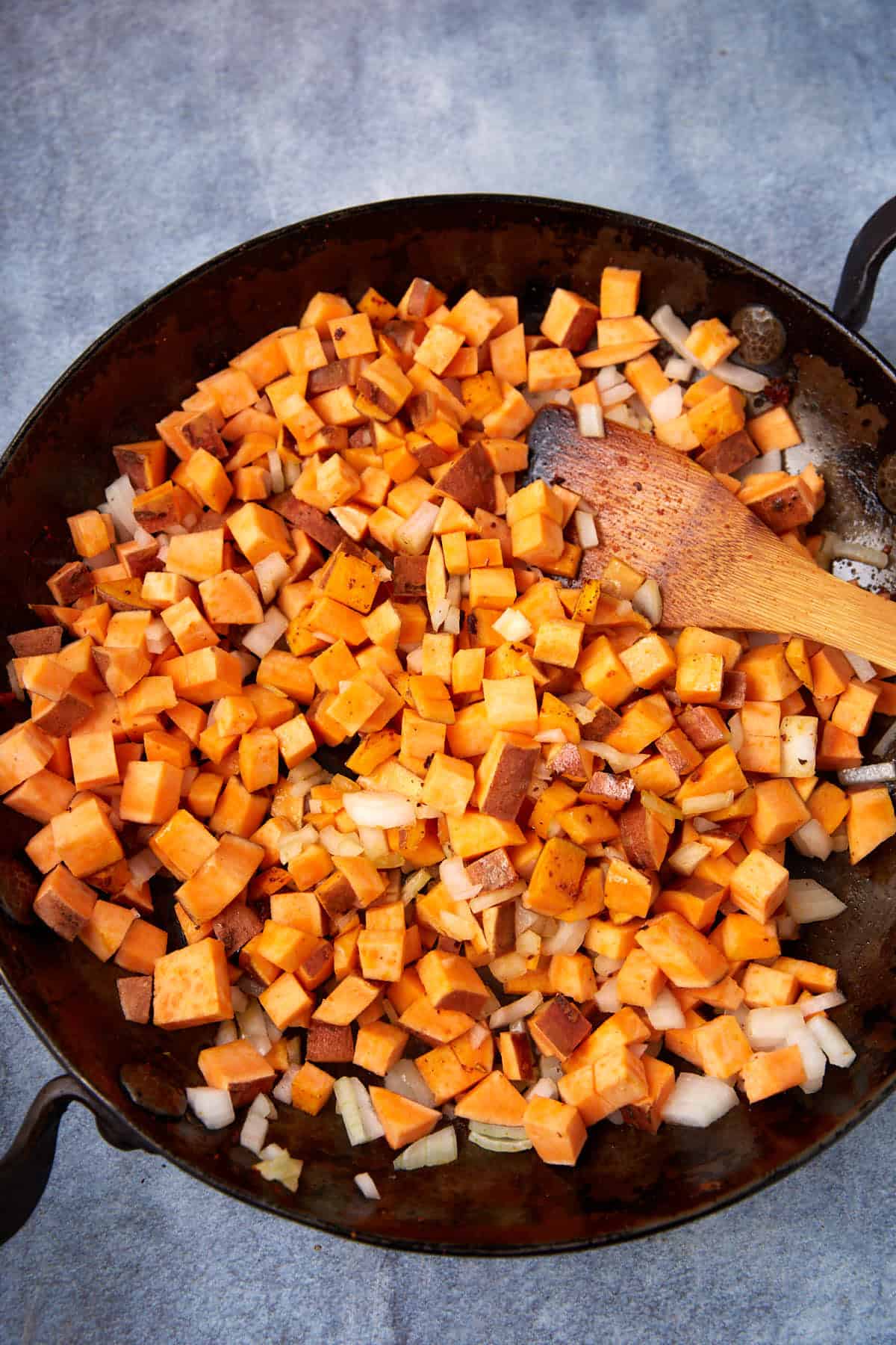 Diced sweet potatoes and chopped onions are being sautéed in a large skillet with a wooden spatula, creating a delicious Sweet Potato Hash, as seen from above.