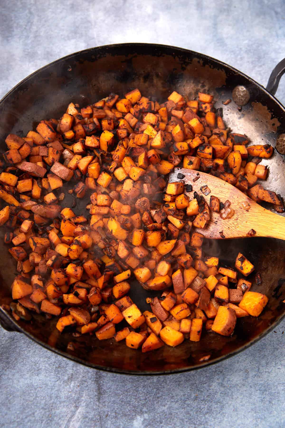Diced, seasoned sweet potatoes sizzle in a large black skillet, stirred with a wooden spoon to create a delicious Sweet Potato Hash. Steam rises from the hot, cooked vegetables against a gray background.