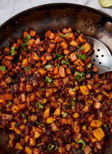 A skillet filled with a savory Sweet Potato Hash made of diced sweet potatoes, onions, and ground meat, topped with chopped fresh herbs, with a metal slotted spoon resting on the side.