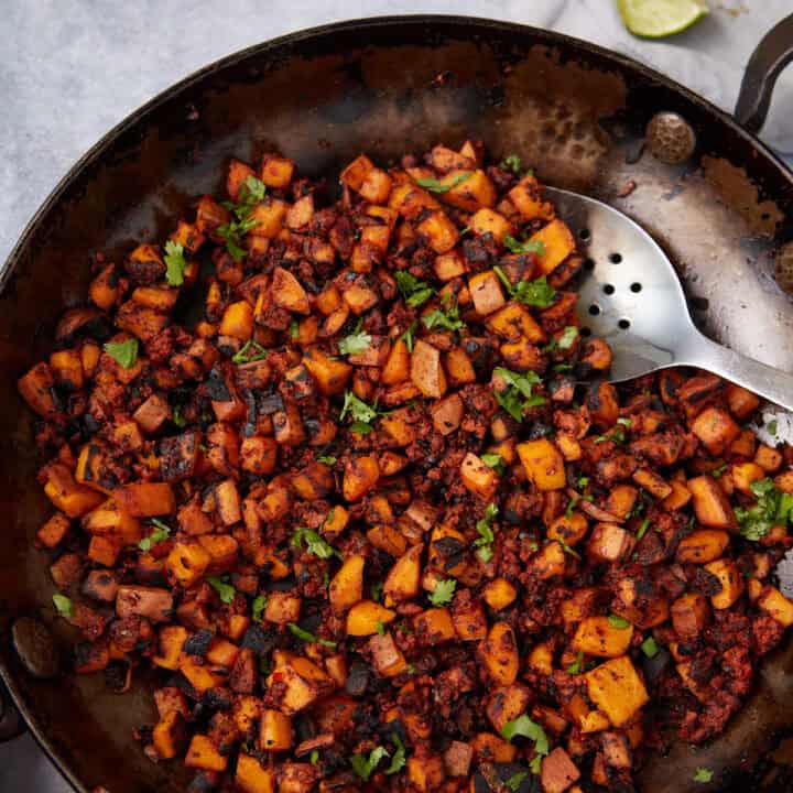 A skillet filled with a savory Sweet Potato Hash made of diced sweet potatoes, onions, and ground meat, topped with chopped fresh herbs, with a metal slotted spoon resting on the side.
