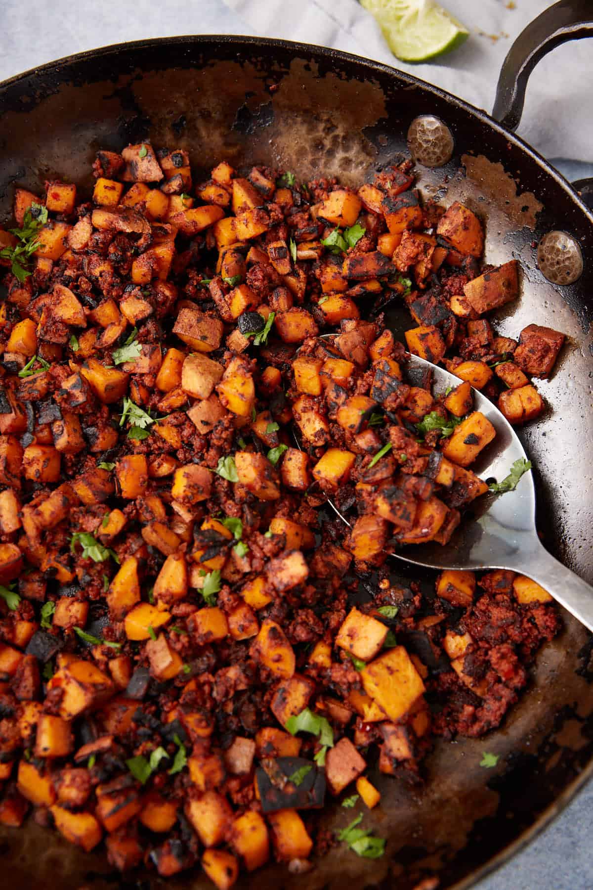 A skillet filled with cooked diced vegetables and crumbled chorizo, garnished with fresh herbs. This vibrant Sweet Potato Hash features orange, brown, and green tones, with a metal spoon resting in the pan, ready to serve.