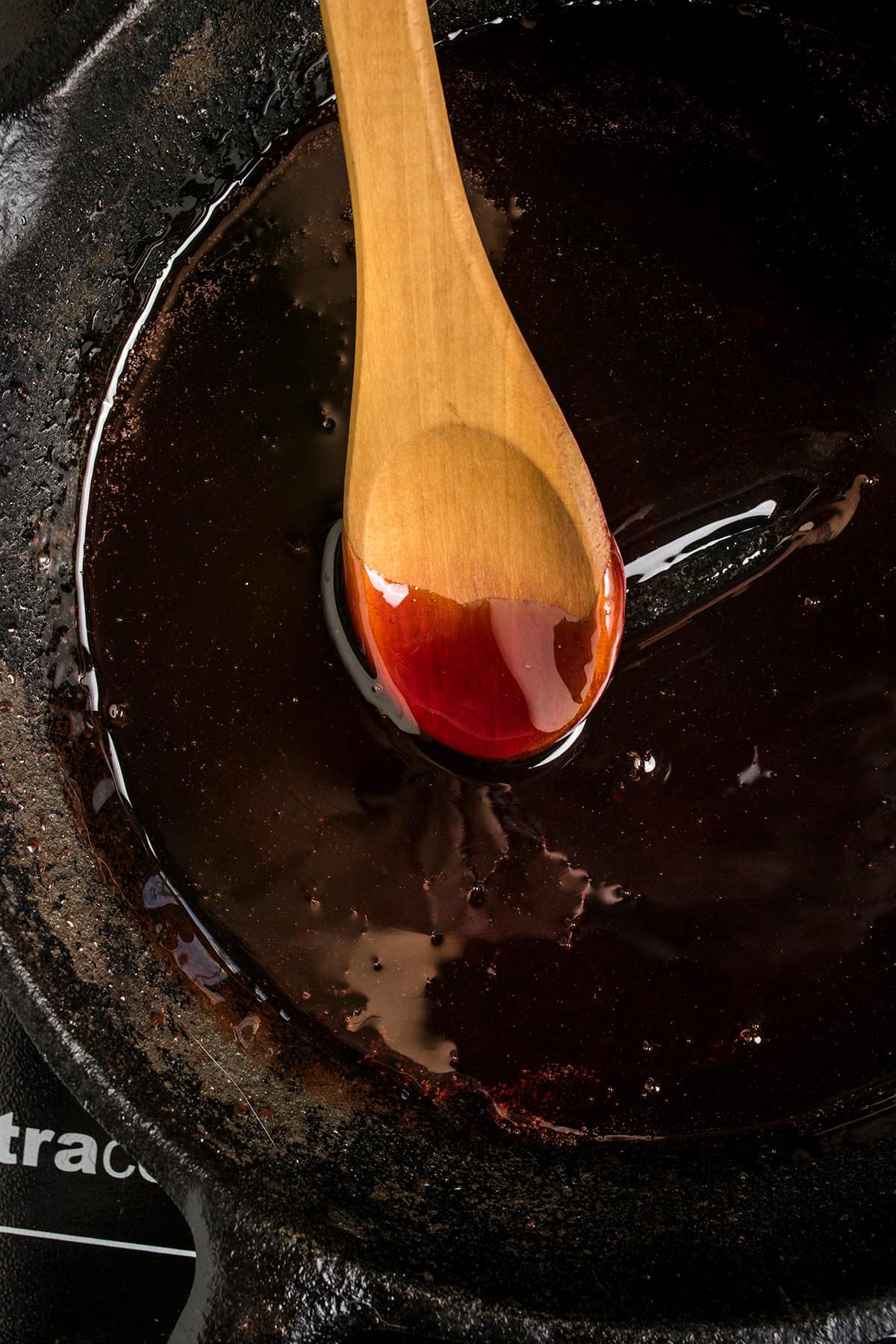 A wooden spoon rests in a cast iron pan, filled with a thick, dark red sauce or glaze, its glossy surface gleaming—perfect for complementing freshly baked Dutch Oven Bread.