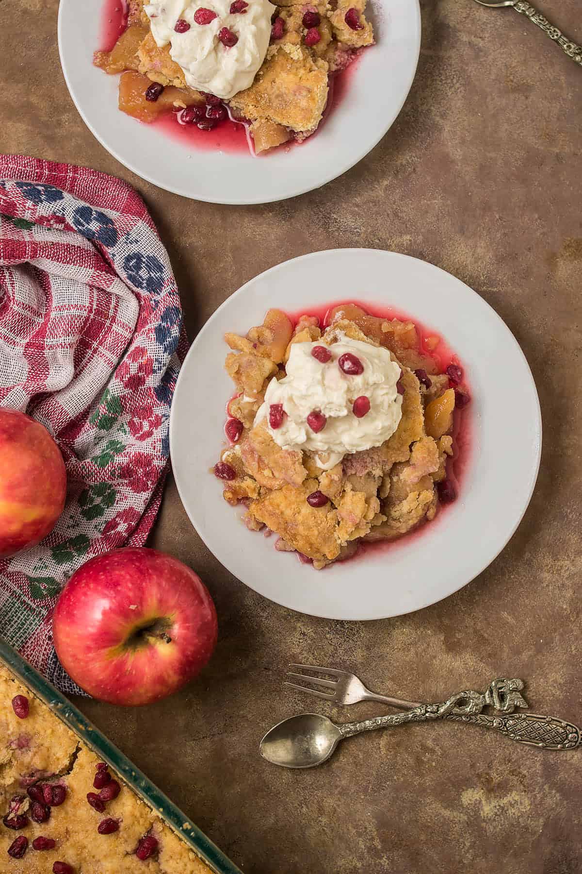 Overhead view of two plates with apple cobbler topped with whipped cream and pomegranate seeds, a red apple, a vintage spoon, Dutch Oven Bread, and a patterned towel on a brown surface.