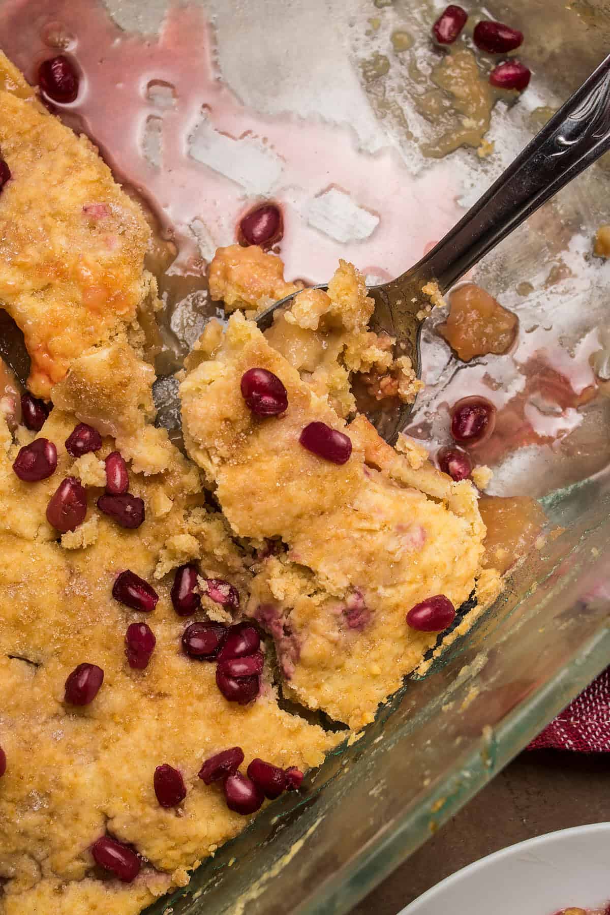 A close-up of a partially eaten dessert in a glass baking dish, topped with pomegranate seeds and served with a metal spoon. Next to the dish sits a slice of golden Dutch Oven Bread, perfect for soaking up the red juice.