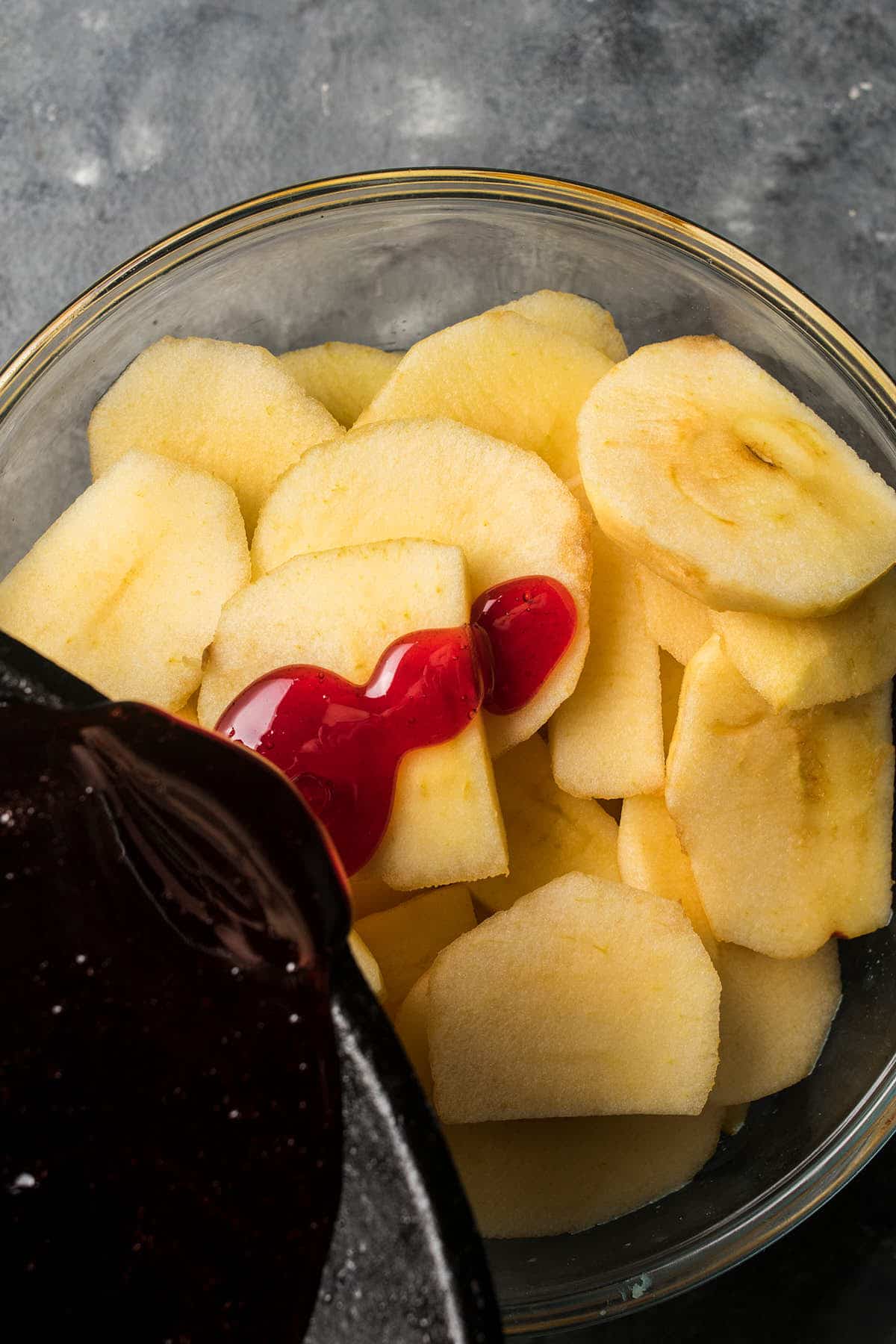 A glass bowl filled with sliced apples, with a thick red sauce being poured over them from a dark pan sits on a gray, textured surface—perfect alongside freshly baked Dutch Oven Bread.
