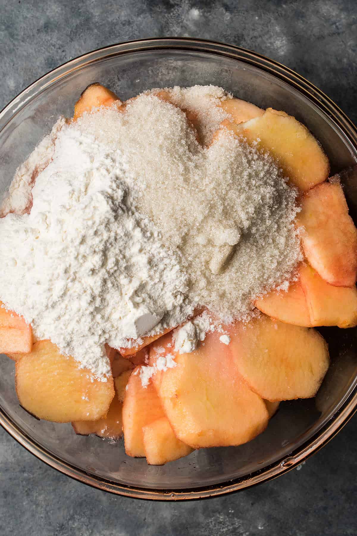 A glass bowl filled with peeled, sliced apples topped with a mound of white flour and granulated sugar, set on a dark gray countertop—perfect inspiration for pairing with warm Dutch Oven Bread.