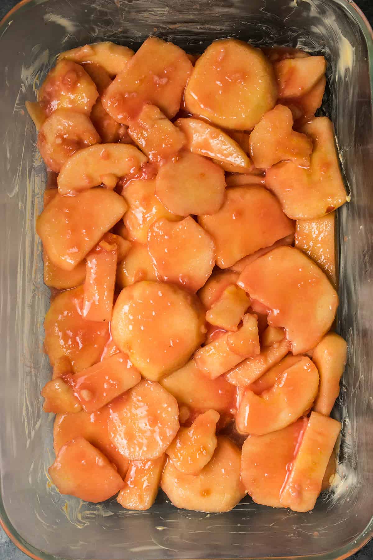 Sliced apples coated in a reddish-orange sauce arranged in a glass baking dish, perfect to enjoy as a sweet side with homemade Dutch Oven Bread.