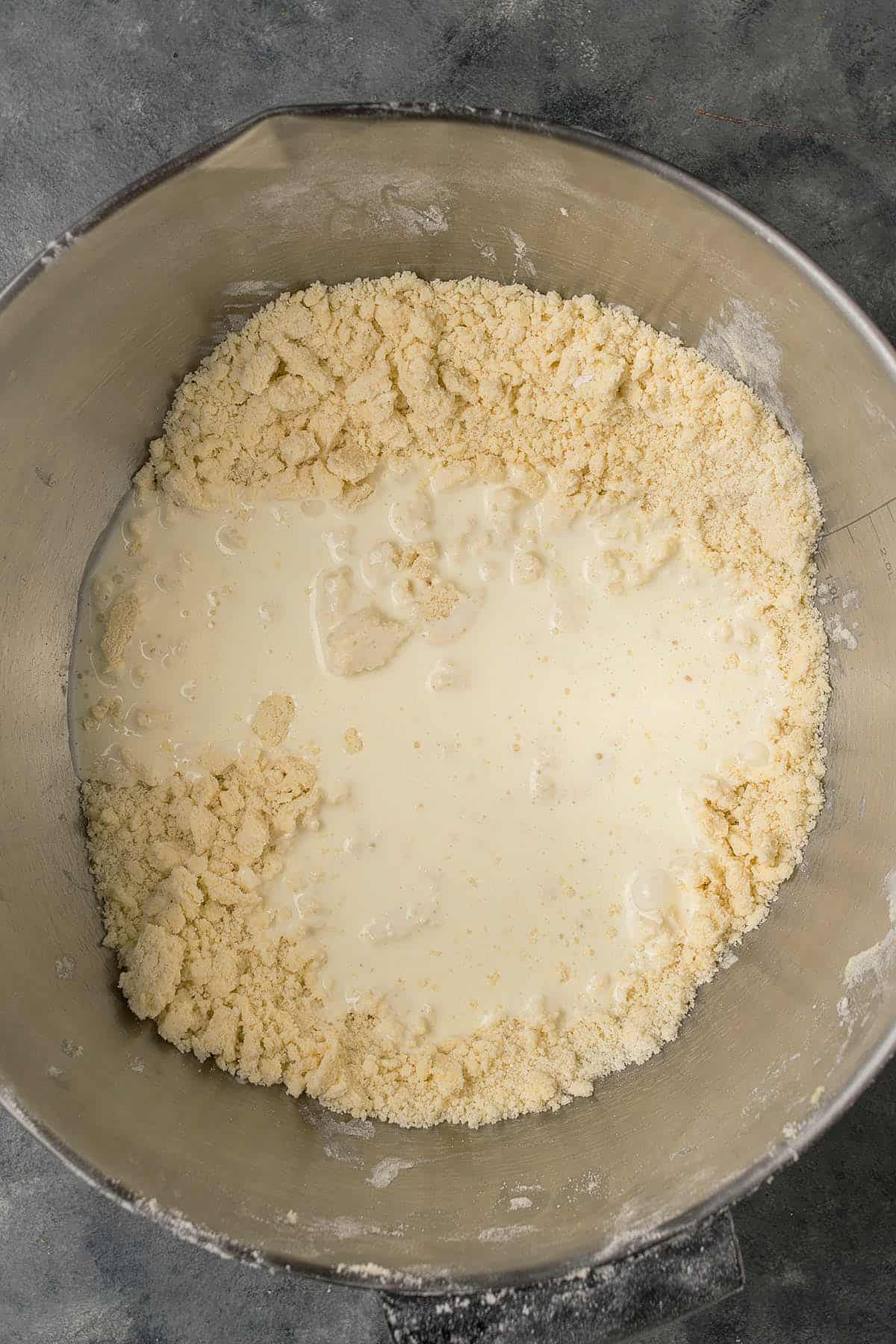 A metal mixing bowl contains crumbly dough with milk or cream poured into the center, ready to be mixed for Dutch Oven Bread on a gray countertop.