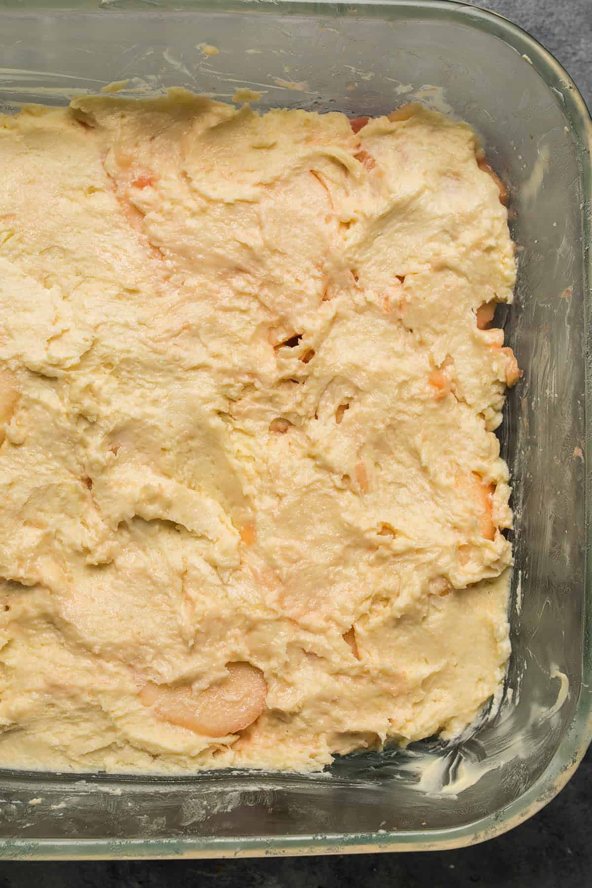 A close-up of unbaked cake batter spread evenly in a glass baking dish, ready to be baked. The creamy, slightly textured surface features orange and yellow tones, similar to the rustic appeal of Dutch Oven Bread dough before baking.