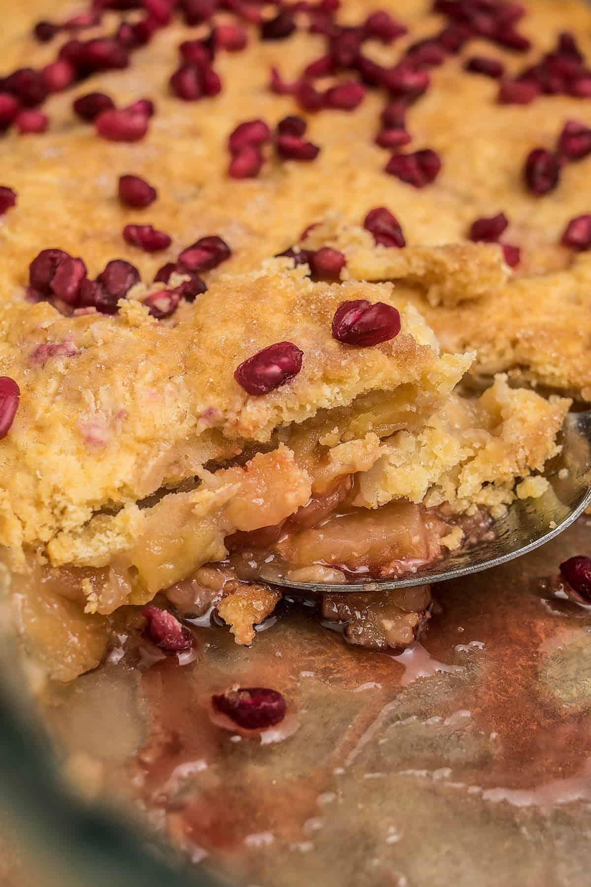 Close-up of a baked fruit cobbler with a golden crust, filled with juicy fruit and topped with pomegranate seeds. Beside it, slices of rustic Dutch Oven Bread complete the scene, as a spoon scoops out the gooey filling inside.