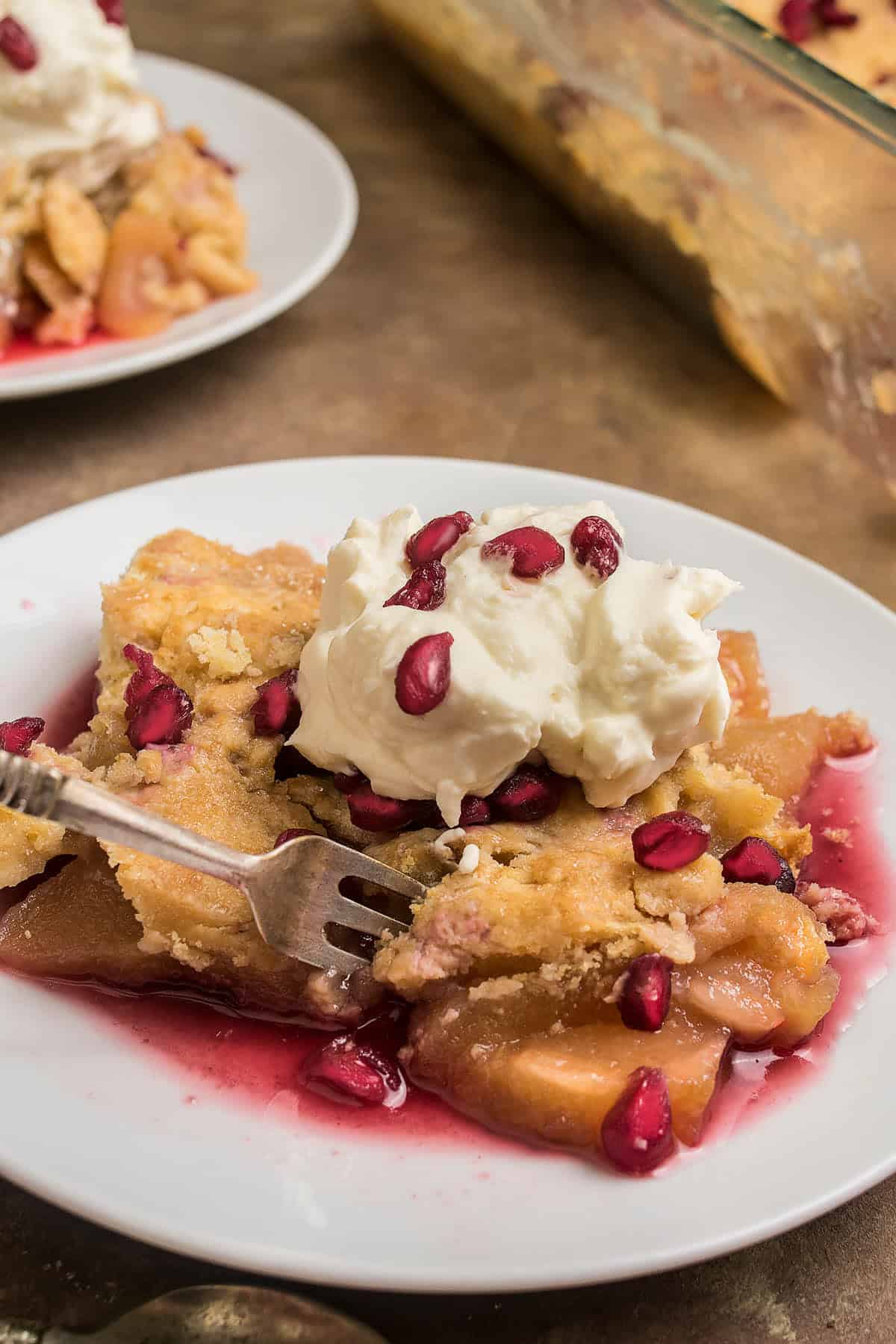 A slice of fruit cobbler topped with whipped cream and pomegranate seeds sits on a white plate beside a fork and glistening fruit juices, while Dutch Oven Bread rests on another plate next to the baking dish in the background.