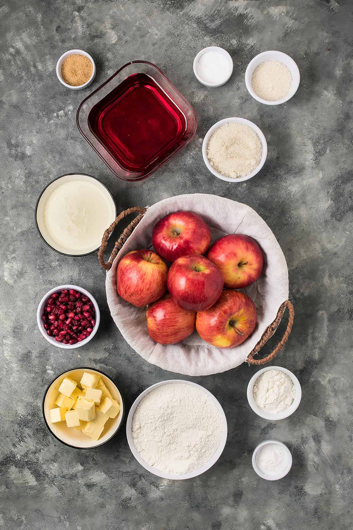 Overhead view of baking ingredients on a gray surface, including a basket of red apples, bowls of flour for Dutch Oven Bread, butter, pomegranate seeds, sugar, spices, a red liquid, and other dry and wet ingredients.