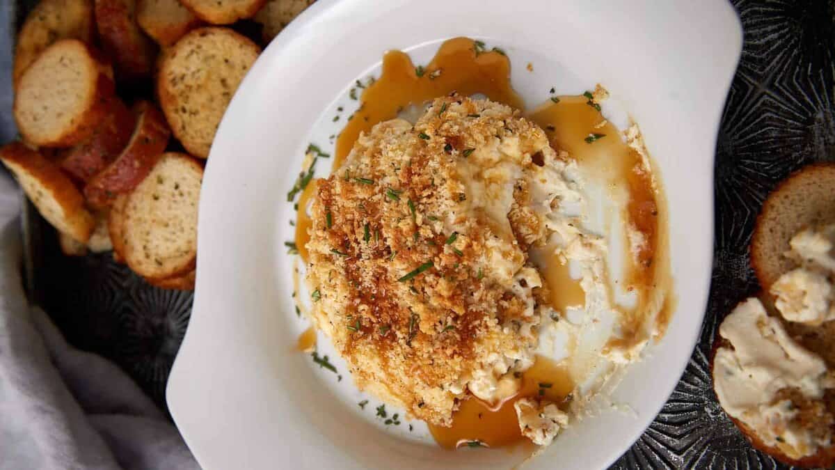 A white dish holds a creamy, baked cheese spread topped with golden breadcrumbs and herbs, with some spread on a toasted baguette slice nearby; more toasted bread slices are in the background.