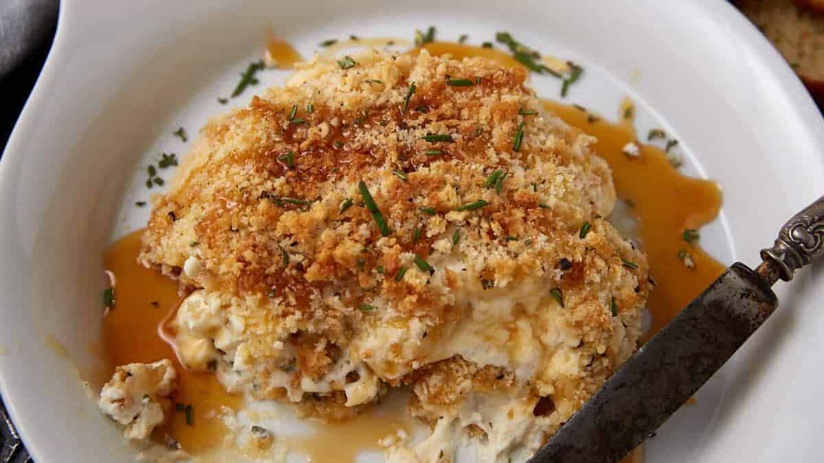 A white dish holds a creamy, baked cheese spread topped with golden breadcrumbs and herbs, with some