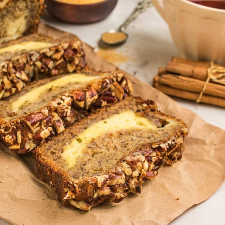 Sliced banana bread with a creamy cheese swirl, topped with chopped pecans, sits on brown parchment paper next to cinnamon sticks and a bowl of brown sugar—perfect alongside homemade Dutch Oven Bread.