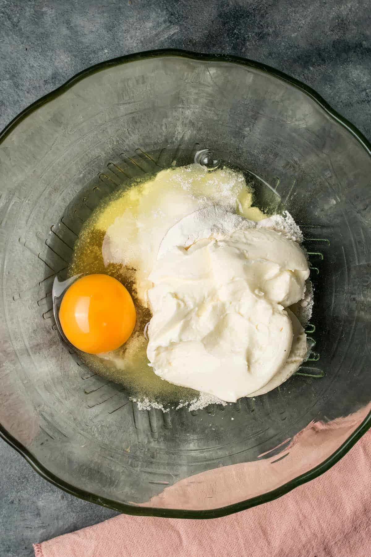 A glass bowl contains an egg, sugar, and ricotta cheese on a gray surface, with a hint of pink cloth at the bottom edge—perfect ingredients to complement your homemade Dutch Oven Bread.