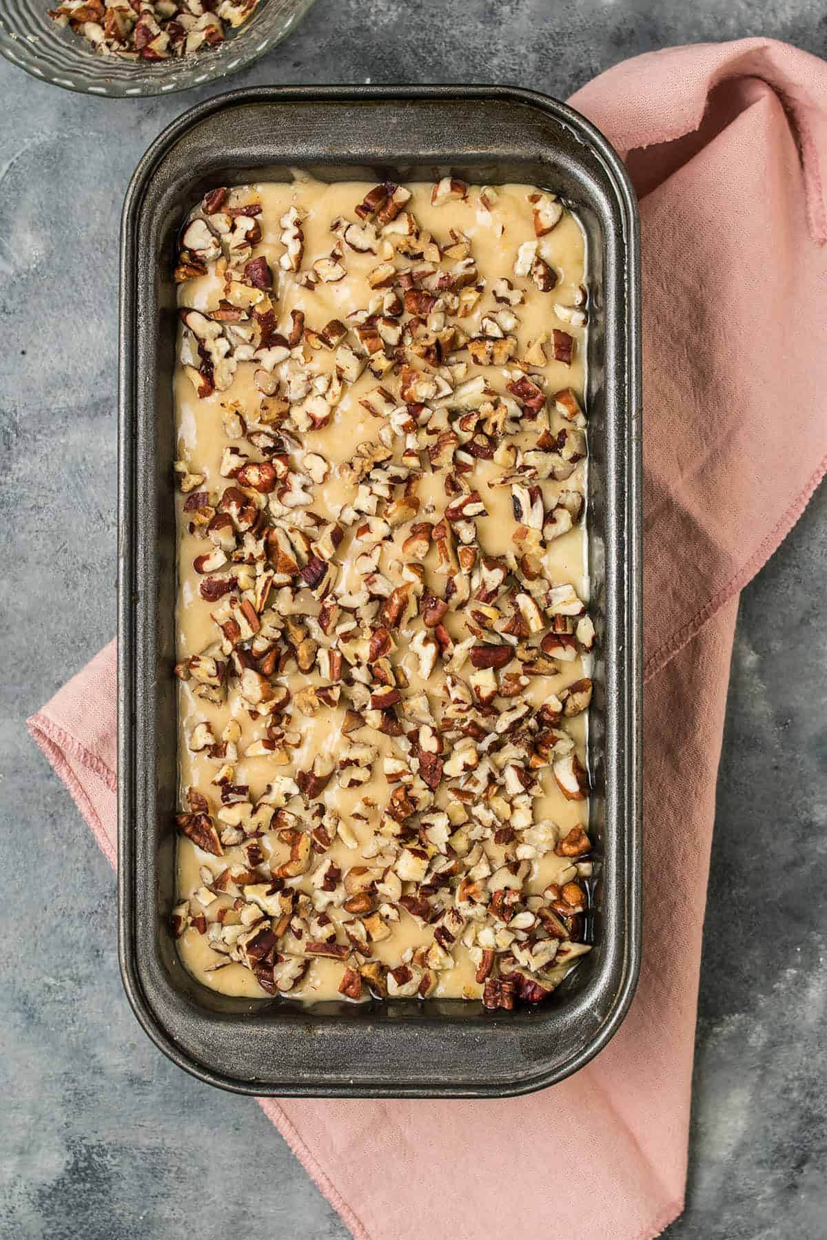 A metal loaf pan filled with unbaked batter topped with chopped pecans, placed on a pink cloth on a gray surface.