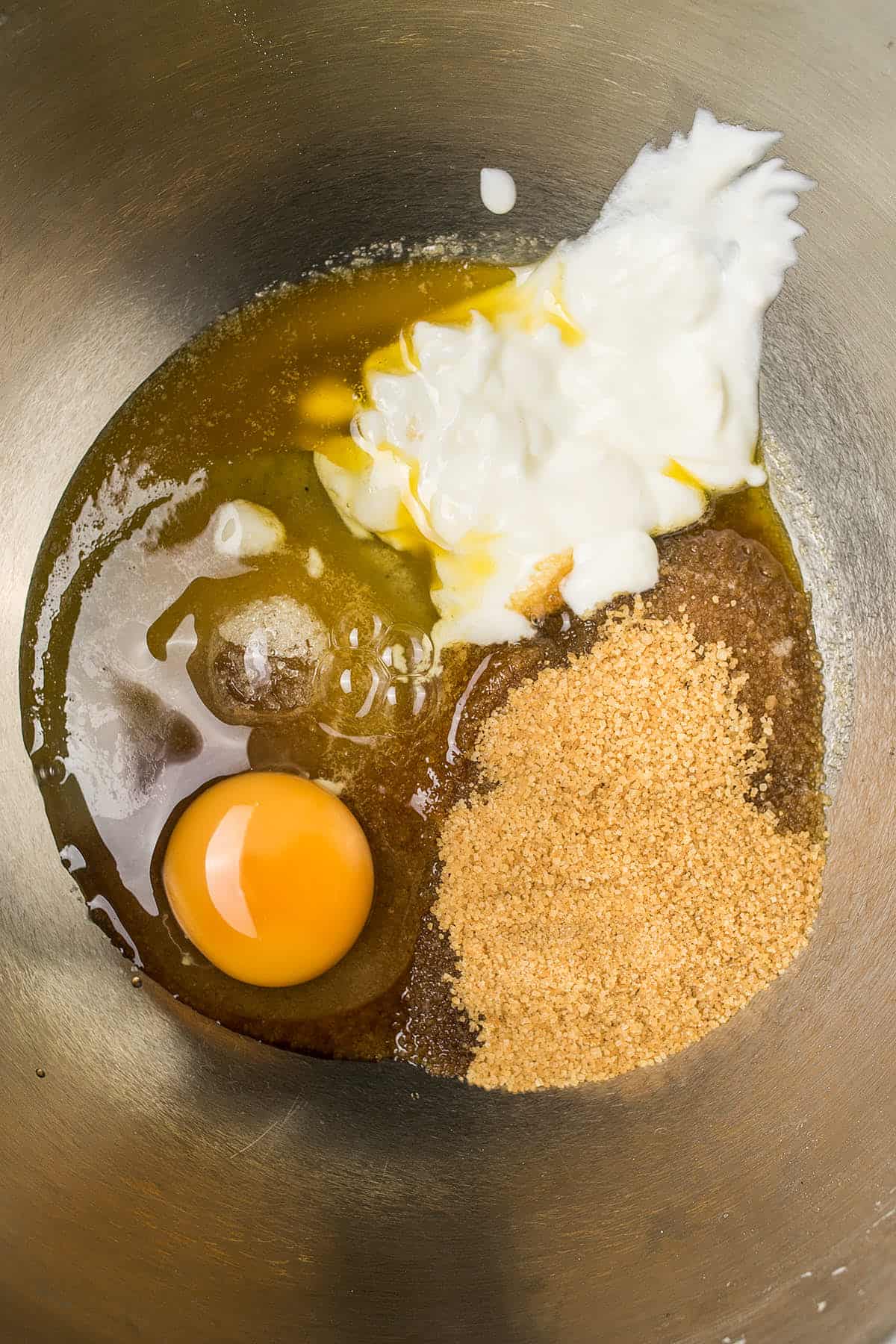 A close-up of a mixing bowl containing ingredients for Dutch Oven Bread—raw egg, brown sugar, granulated sugar, yogurt, and melted butter—all unmixed and arranged in separate sections.
