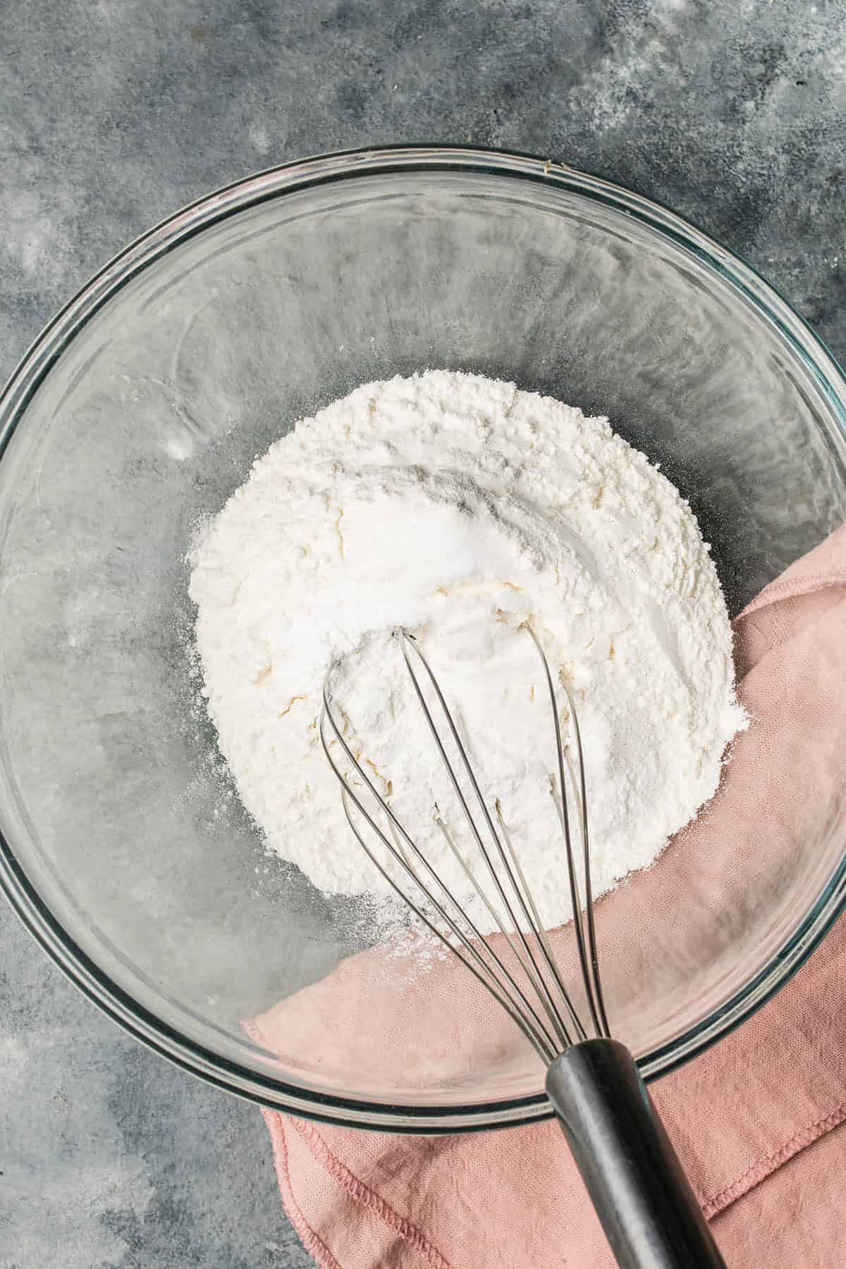 A glass mixing bowl with flour and a metal whisk inside, ready to prepare Dutch Oven Bread, rests on a pink cloth atop a gray textured surface.
