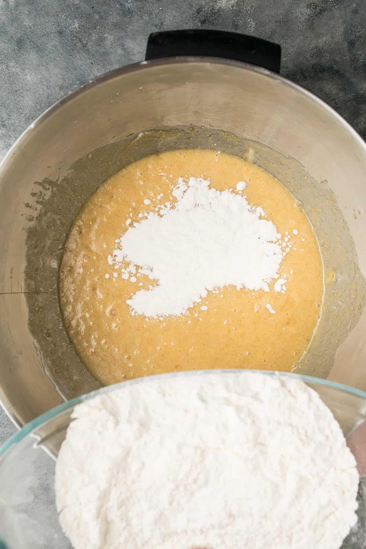 A metal mixing bowl with yellow batter and a mound of white flour on top, as more flour is poured in from a glass bowl above—a key step in preparing dough for Dutch Oven Bread. The scene unfolds on a gray countertop.