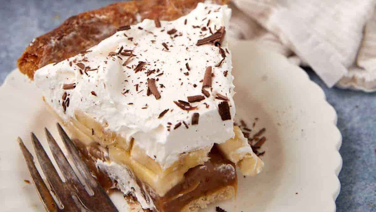 A slice of banoffee pie topped with whipped cream and chocolate shavings sits on a white plate beside a fork—one of those Christmas pies you can't mess up, with visible layers of banana and caramel filling.