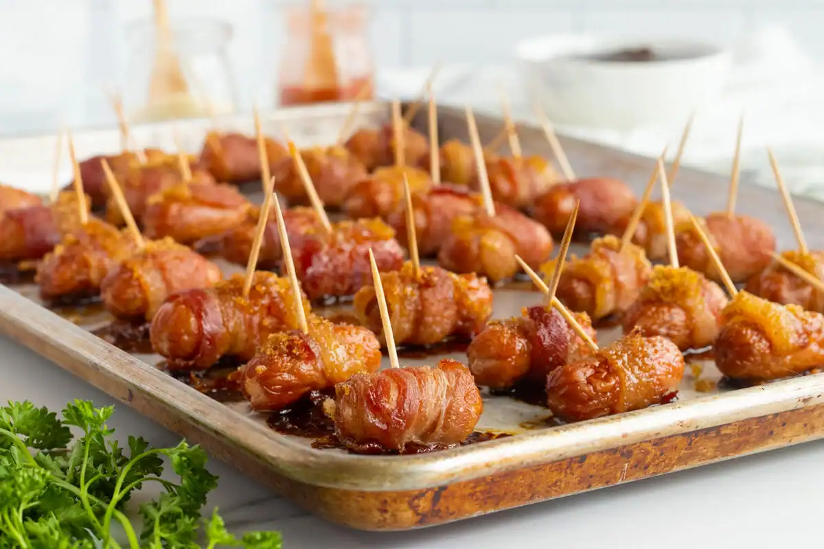 A baking tray filled with bite-sized holiday appetizers—bacon-wrapped sausages held together with toothpicks, sprinkled with brown sugar and baked. Fresh parsley is in the foreground, with blurred bowls and sauce jars in the background.
