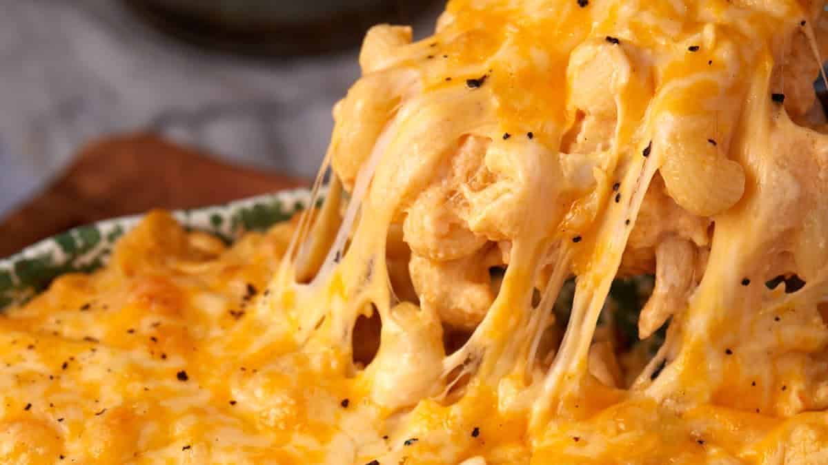 A close-up of buffalo mac and cheese in a casserole dish, with gooey melted cheese stretching as a portion is lifted out, topped with cracked black pepper.