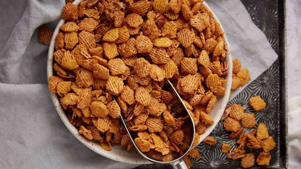 A bowl filled with crunchy cereal pieces, with a metal scoop resting inside. Some cereal is scattered on a tray beside the bowl, all placed on a light-colored cloth.