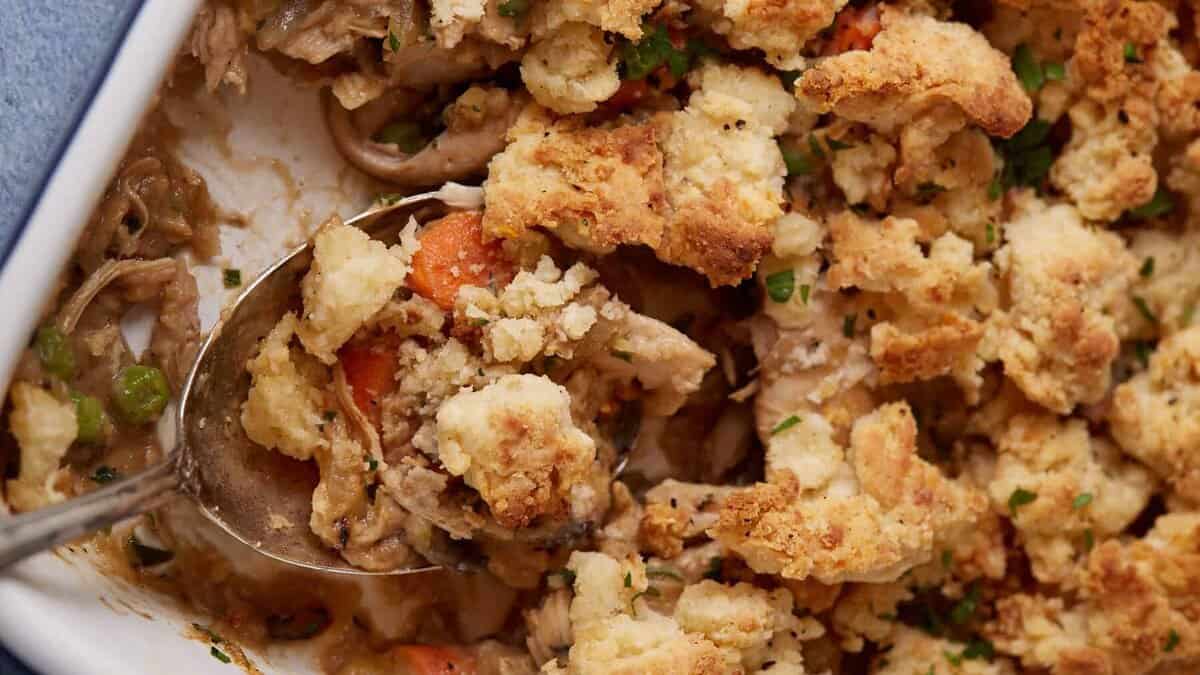 A close-up of a savory casserole is shown. The dish features golden-brown, crispy biscuit topping over a filling of shredded chicken, carrots, peas, and a creamy sauce. A spoon is scooping out some of the casserole, revealing the ingredients beneath the topping.