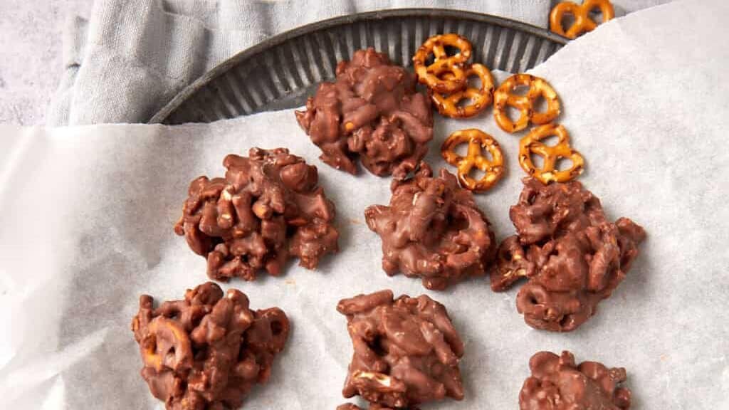 Clusters of chocolate-covered pretzels arranged on parchment paper atop a round metal tray, with a few whole pretzels placed in the corner. The background is a light, textured surface.