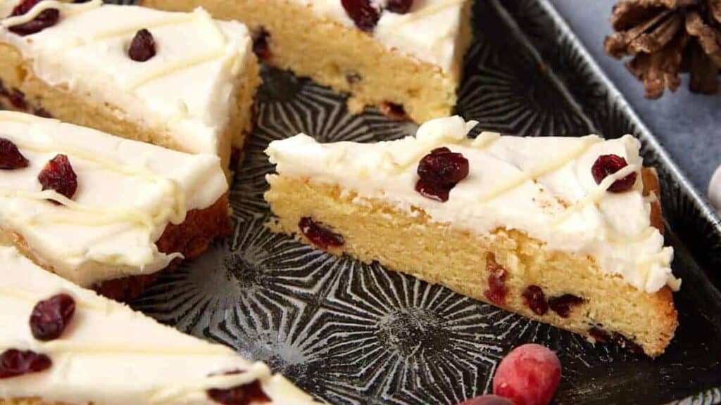 Sliced cranberry bliss bars with white icing and dried cranberries arranged on a dark patterned tray, with two fresh cranberries nearby—these are Christmas bar desserts that vanish faster than cookies.