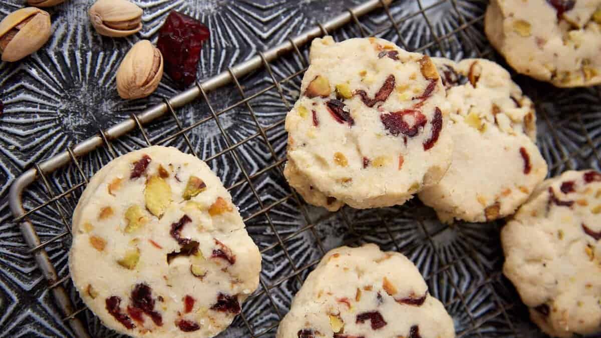 Christmas cookies made for the busiest part of December, these treats with pistachios and dried cranberries are cooling on a metal rack, while whole pistachios and cranberries add festive color to the textured surface nearby.